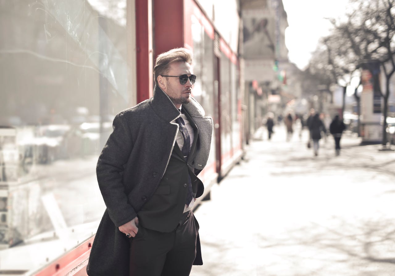 A fashionable man in a coat and sunglasses walking on a sunny city street.